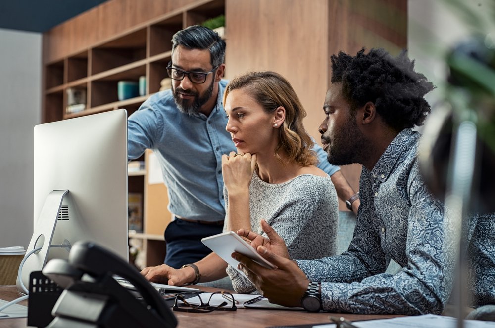 Focused businesspeople discussing project while looking at computer screen. Mature manager working with his creative team in a modern office. Multiethnic casual teamwork at work.