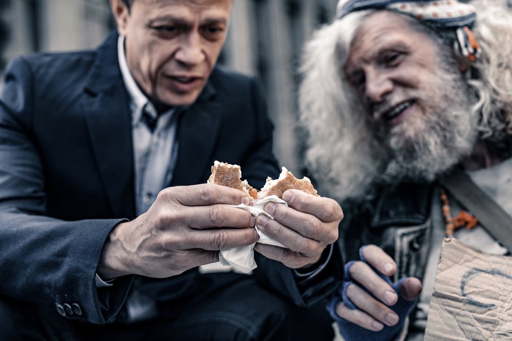 Begging for food. Sincere kind man in office costume sharing sandwich with homeless man while sitting close to him