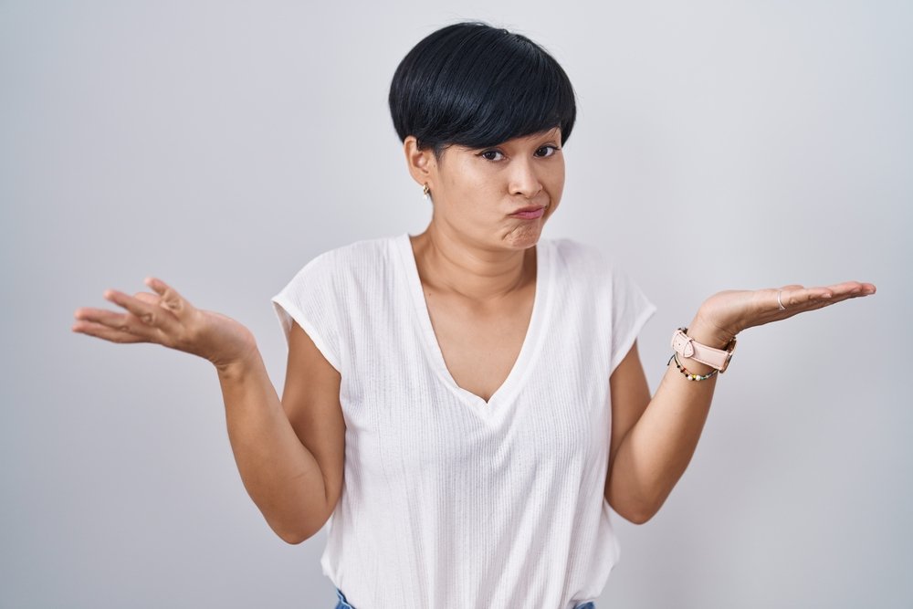 Young asian woman with short hair standing over isolated background clueless and confused expression with arms and hands raised. doubt concept.