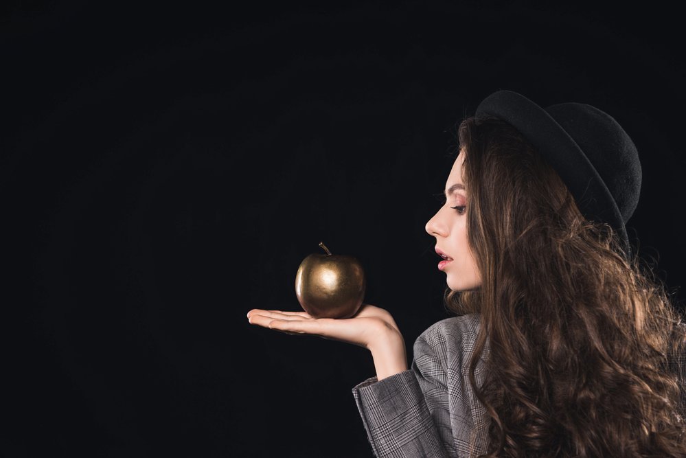 Side view of fashionable young woman holding golden apple on palm isolated on black