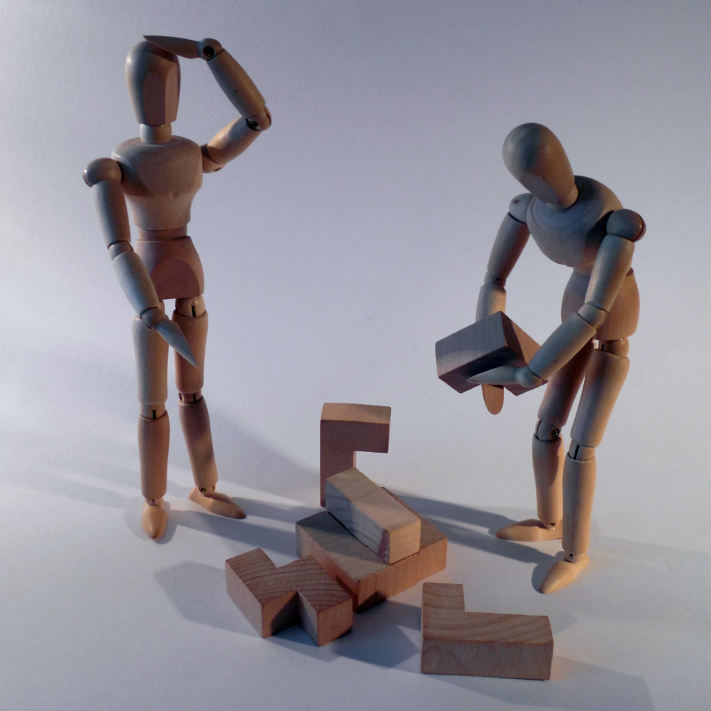 two wooden figures standing, one of them lifting up bricks, other is just looking with one hand on his head