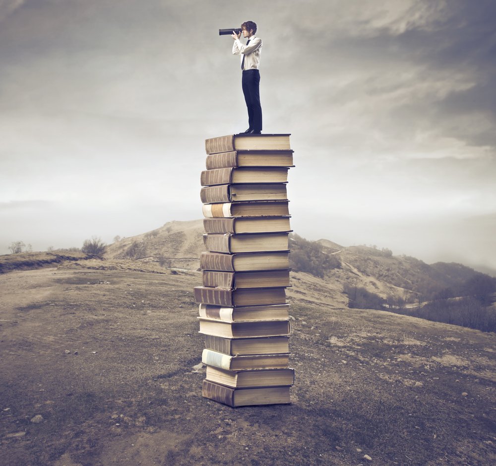 man standing on long pile of books looking through binoculars